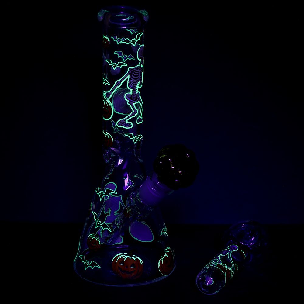 A glowing glass water pipe with a black base and neon green Halloween-themed designs, shown in a dark room with a separate glass mouthpiece on the right.